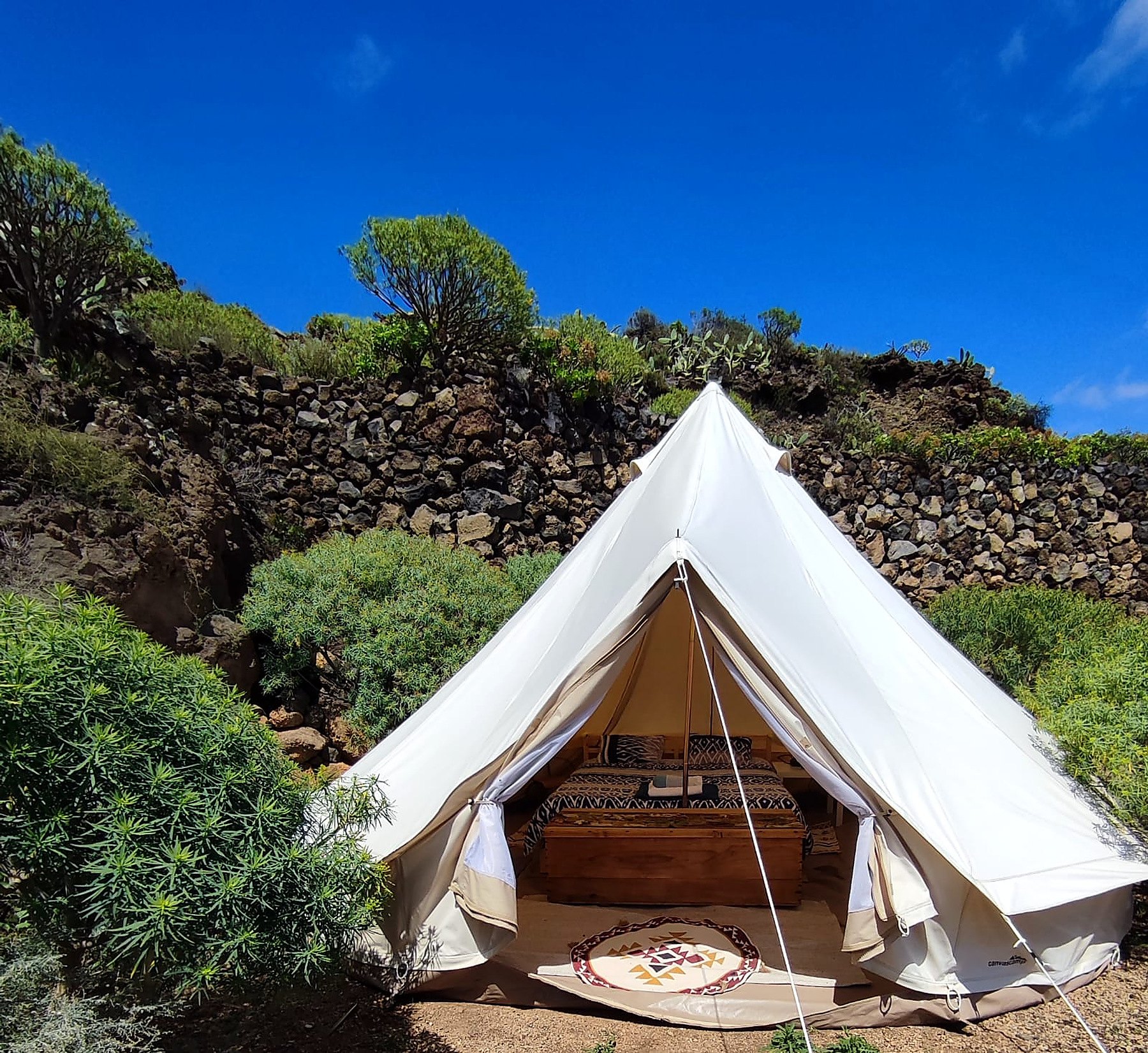Tente de glamping blanche ouverte sur un lit, adossée à un mur de pierres volcaniques et à la végétation